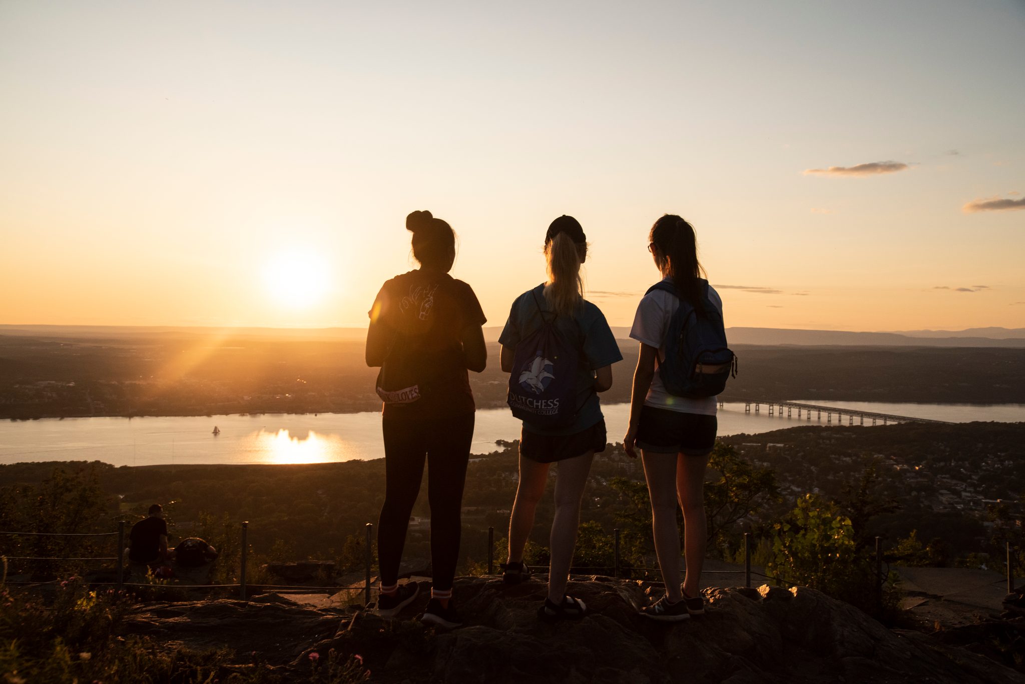 Three hikers stand on the ledge as the sun drops toward the river, the bridge a thin line of light below.