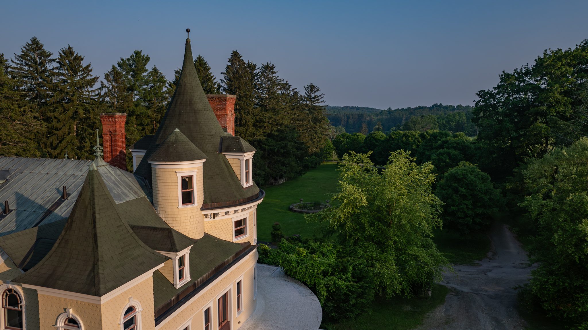 Last warm light on the turrets and shingles, the drive curving away into trees that are already in shadow.