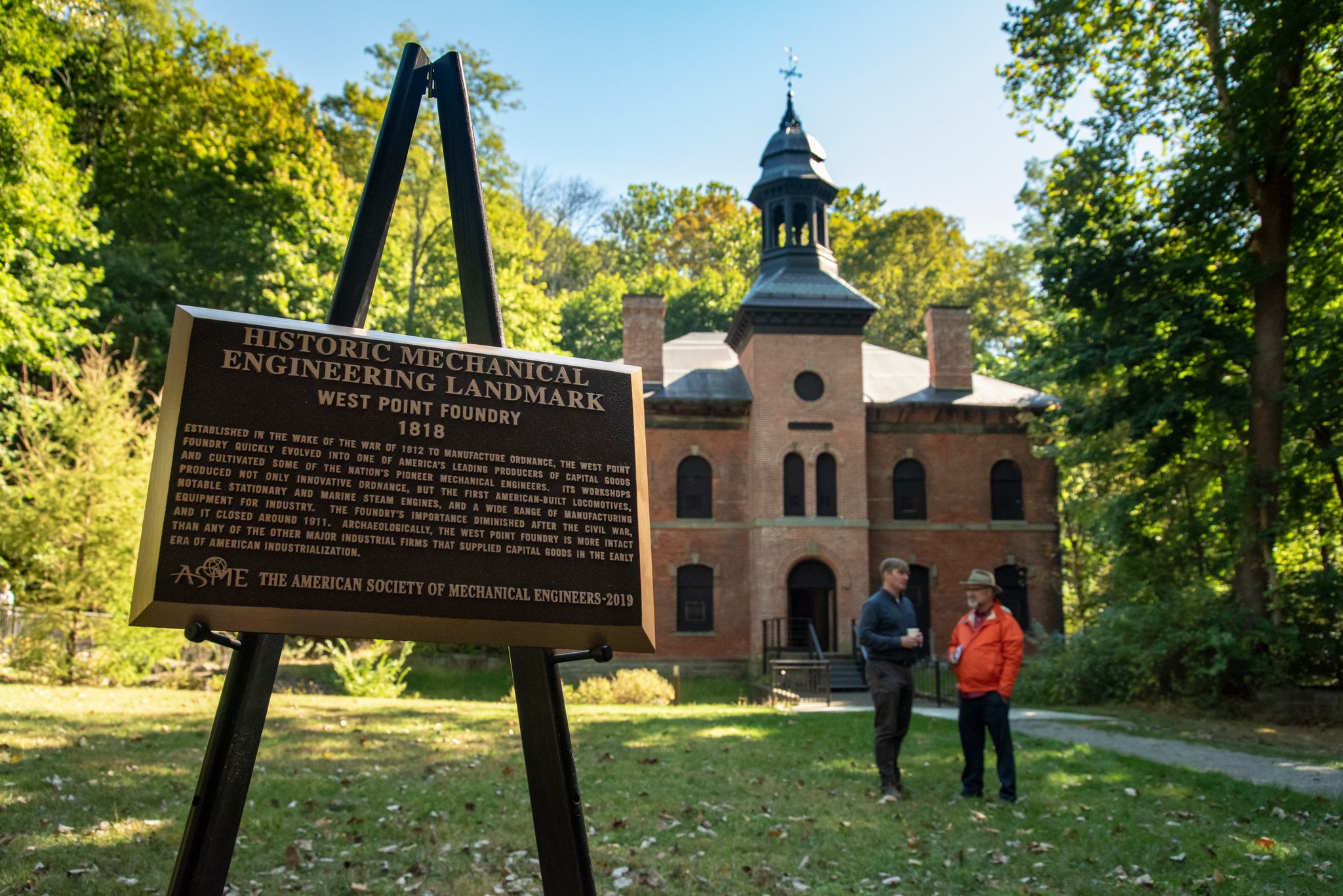 A plaque names the West Point Foundry a historic engineering landmark, its brick building and cupola standing behind.