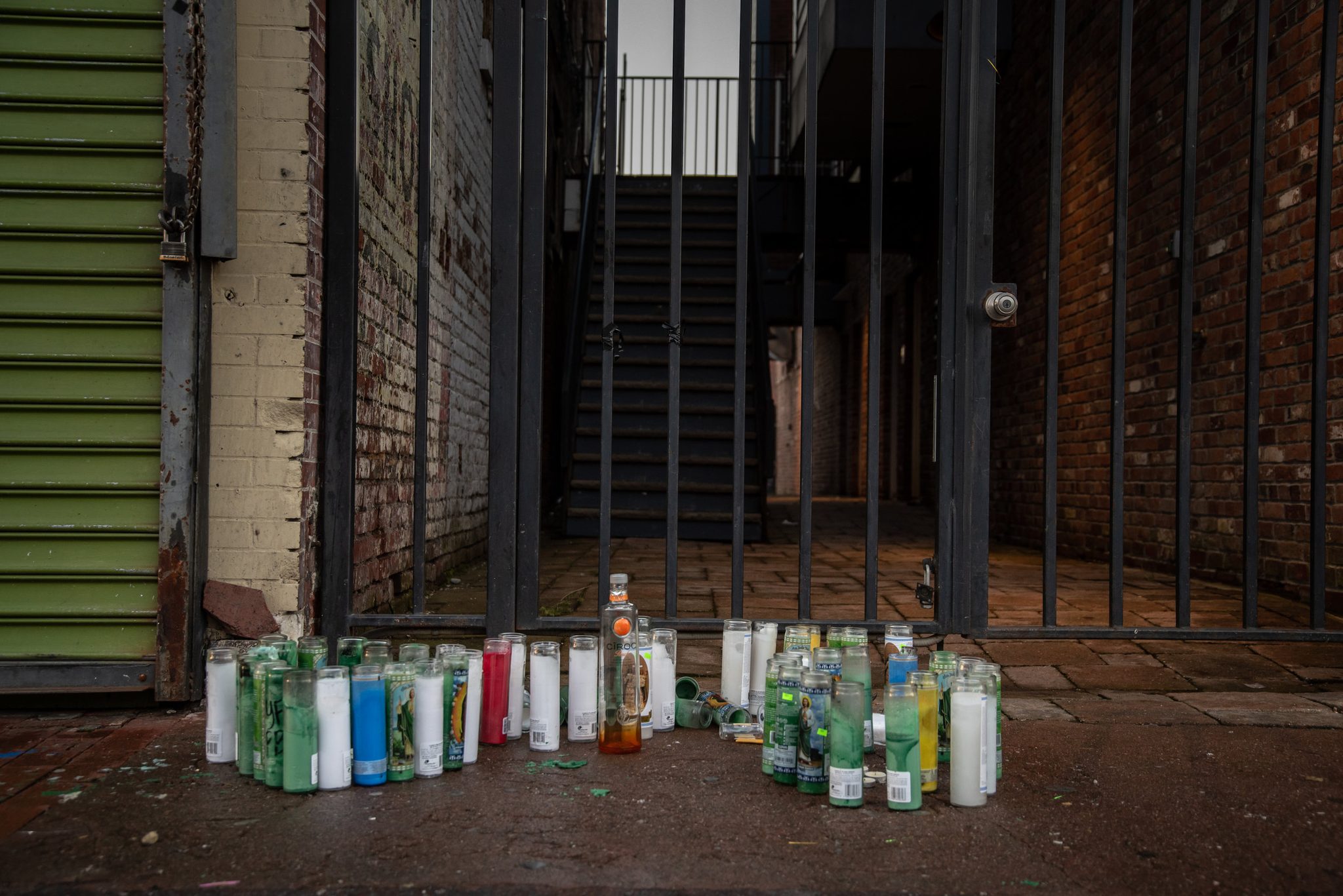 Candles crowd the sidewalk in front of a locked gate, a memorial left for someone.