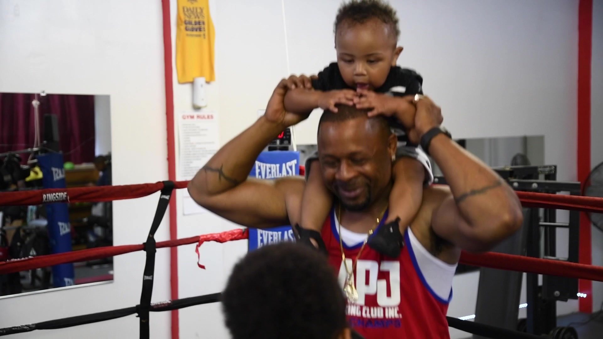 Coach Kariym grins under a toddler riding on his shoulders, both small hands resting on top of his head.