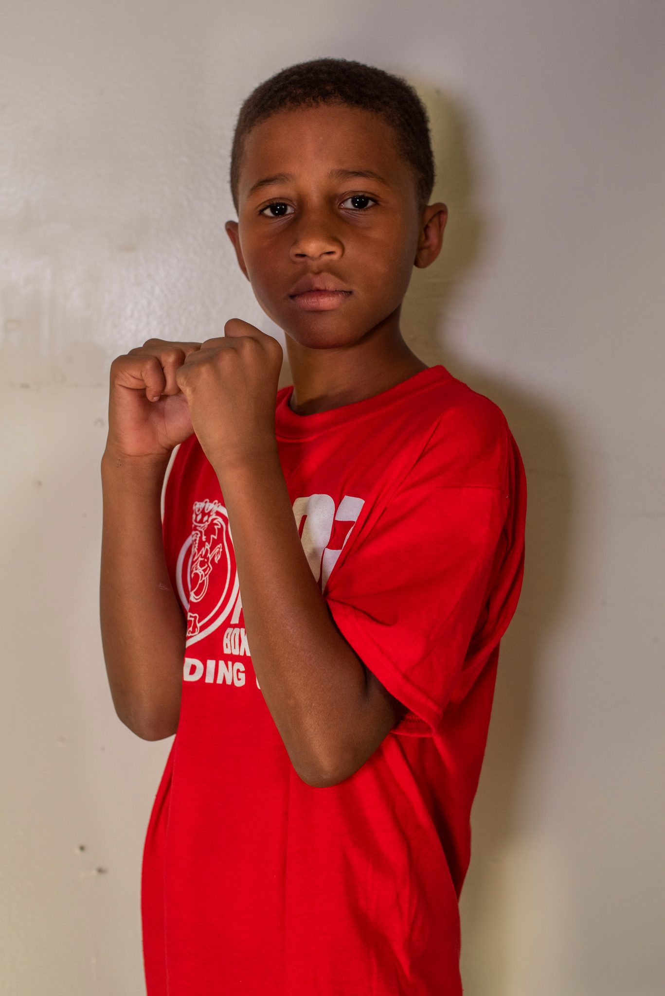 Against the white wall, a young boxer lifts his fists to his chest and meets the lens without blinking.