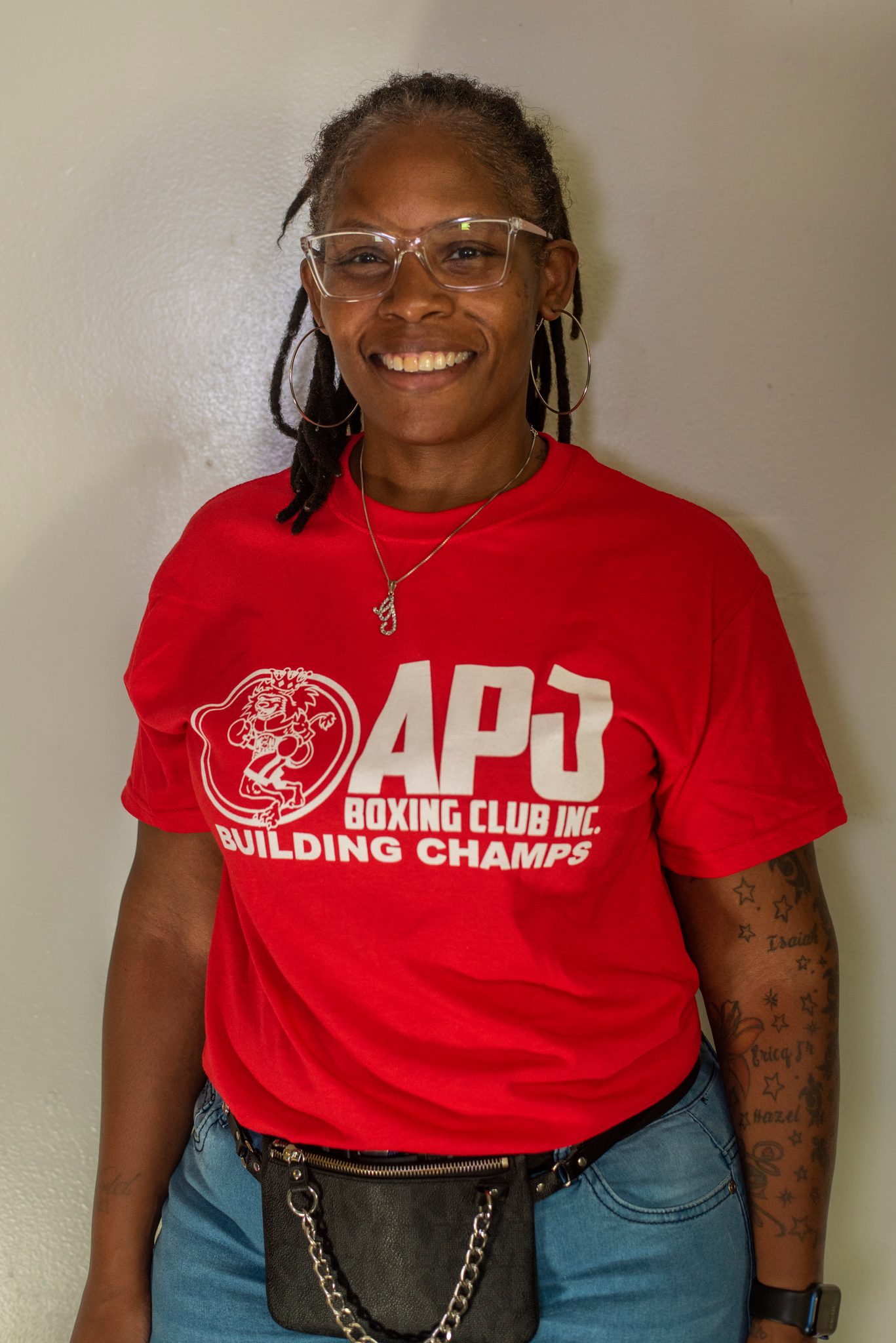 Glasses, long braids and a wide smile, she stands easy in the club shirt with a chain-strap bag at her hip.