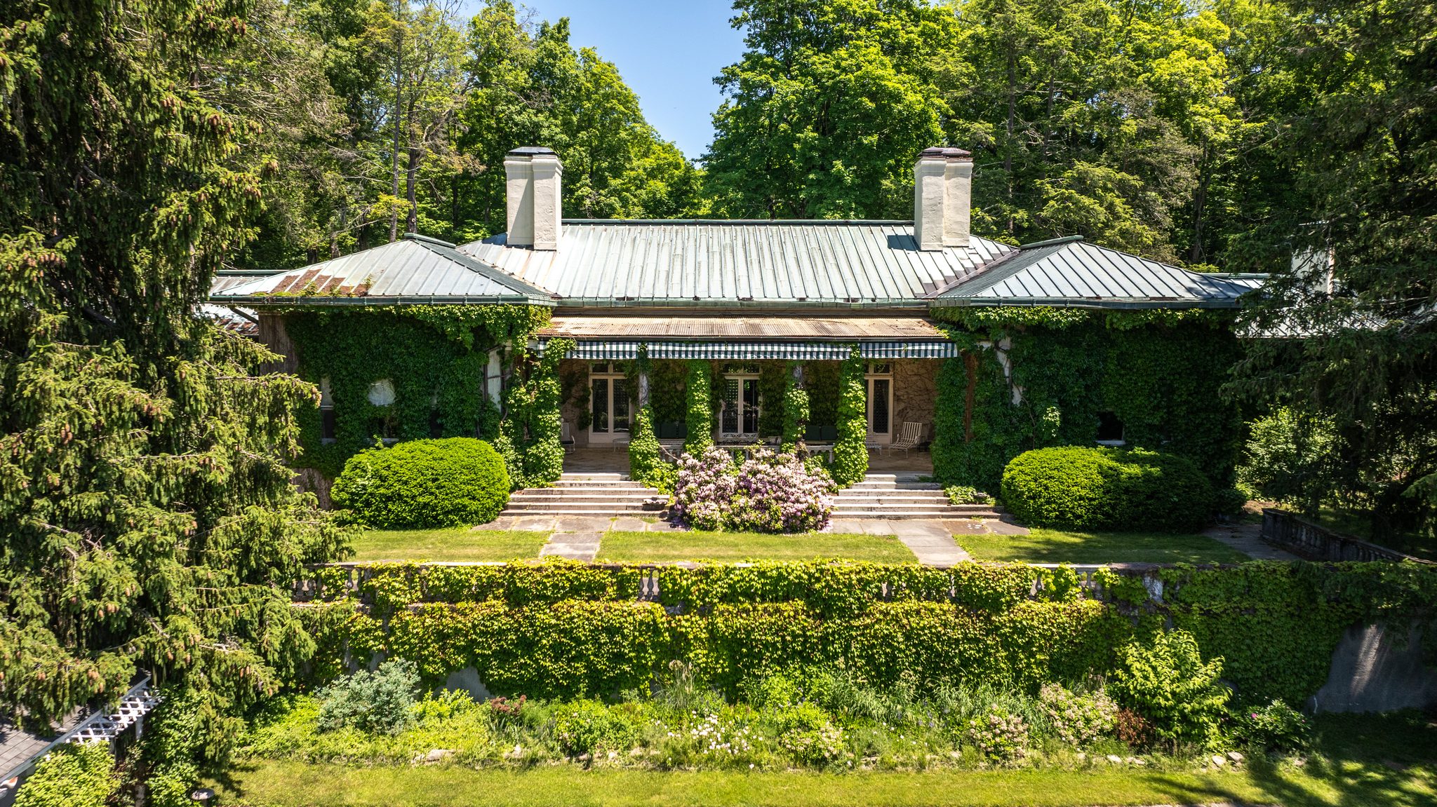 The bungalow's garden front, stone steps rising through clipped hedges and rhododendron to a striped awning and French doors.