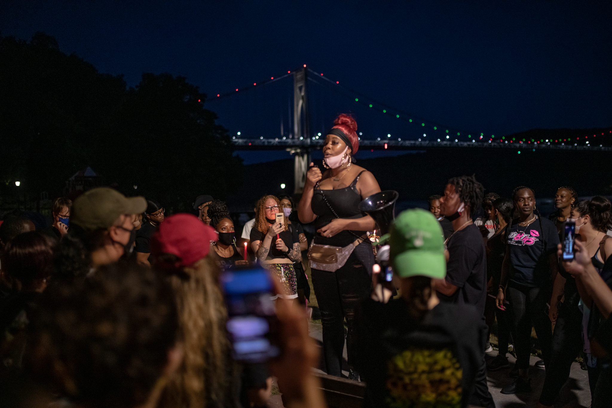 A woman speaks to the crowd by the river, the Mid-Hudson Bridge lit up behind her.