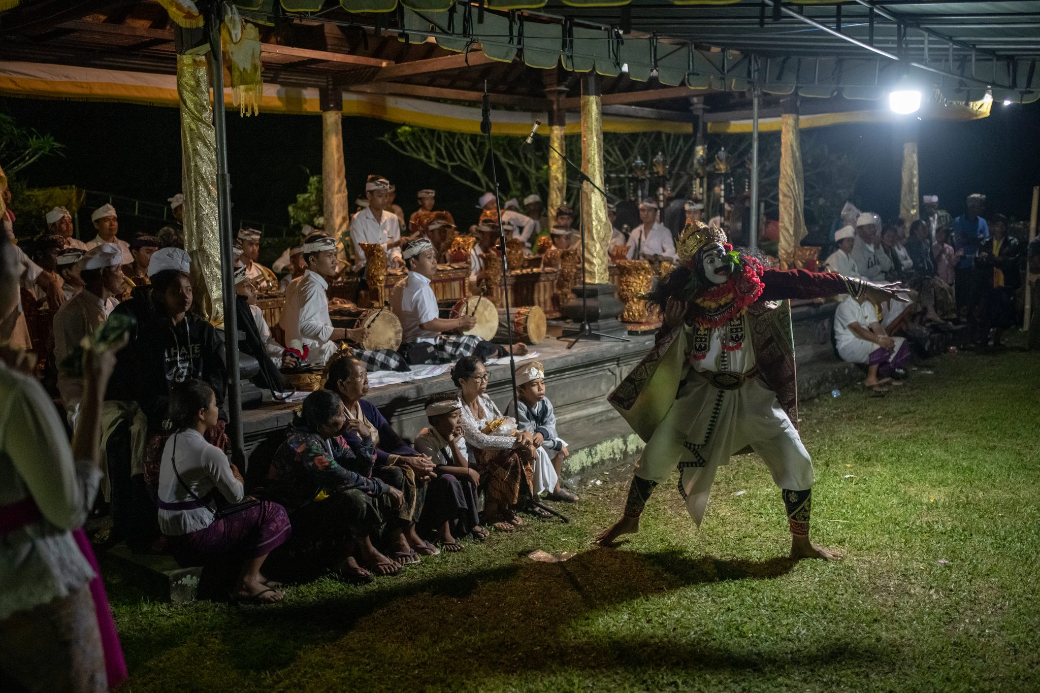 After dark, a masked dancer lunges across the grass while the gamelan plays under the lights.