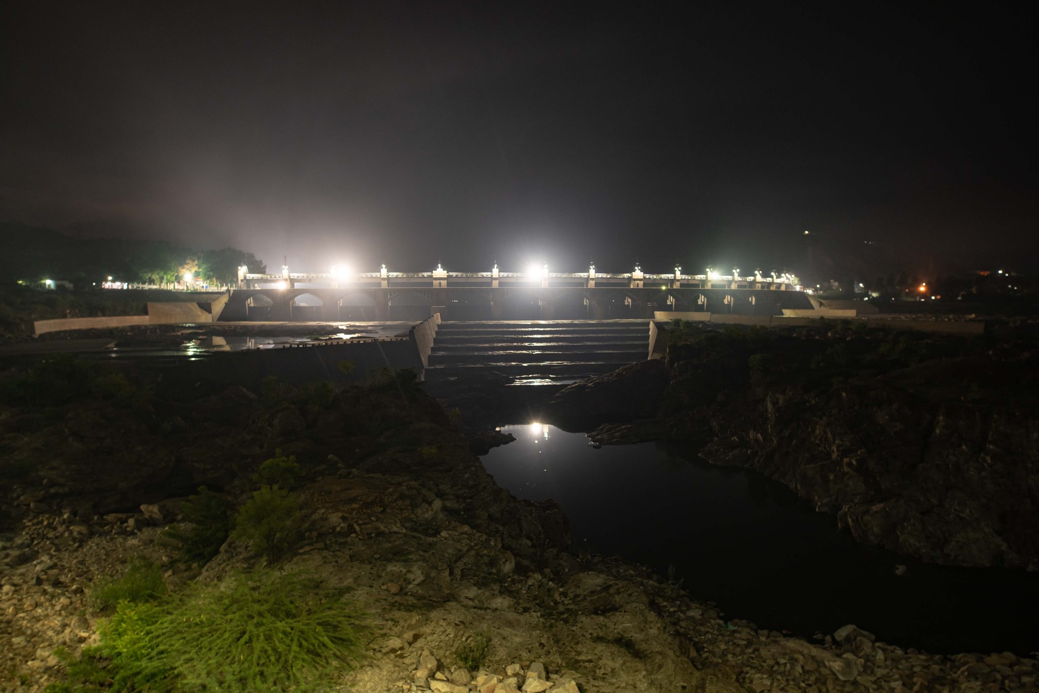 Floodlights blaze along the dam at night, the water black below.