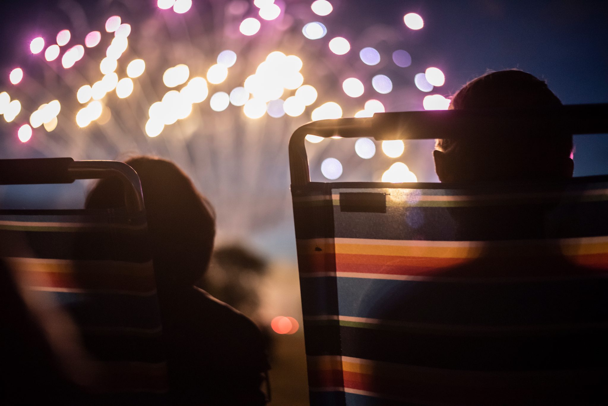 Heads tip back in folding chairs as the fireworks bloom out of focus overhead.