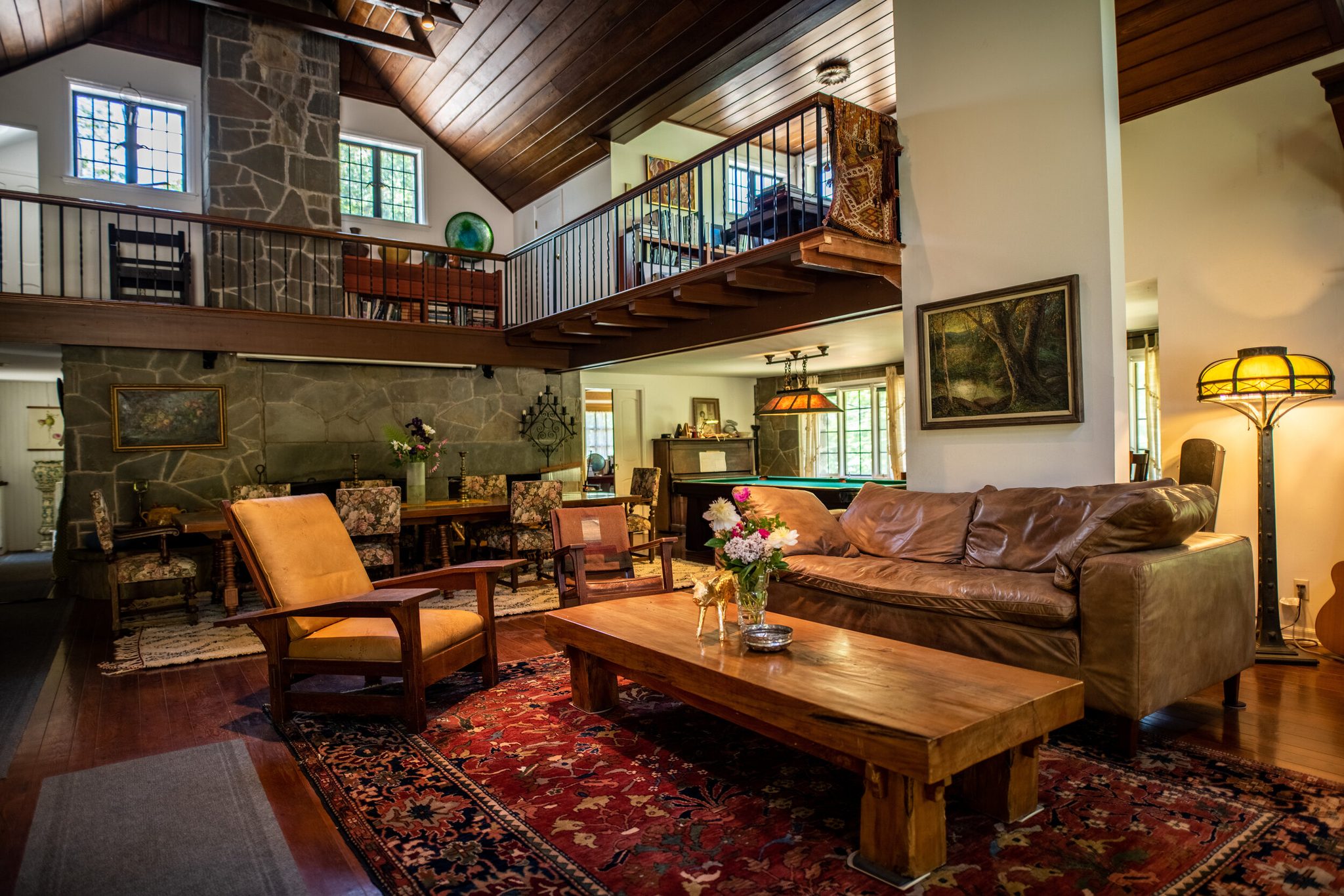 A stone chimney rises to a dark plank ceiling in this June great room, with a gallery above and warm lamps below.