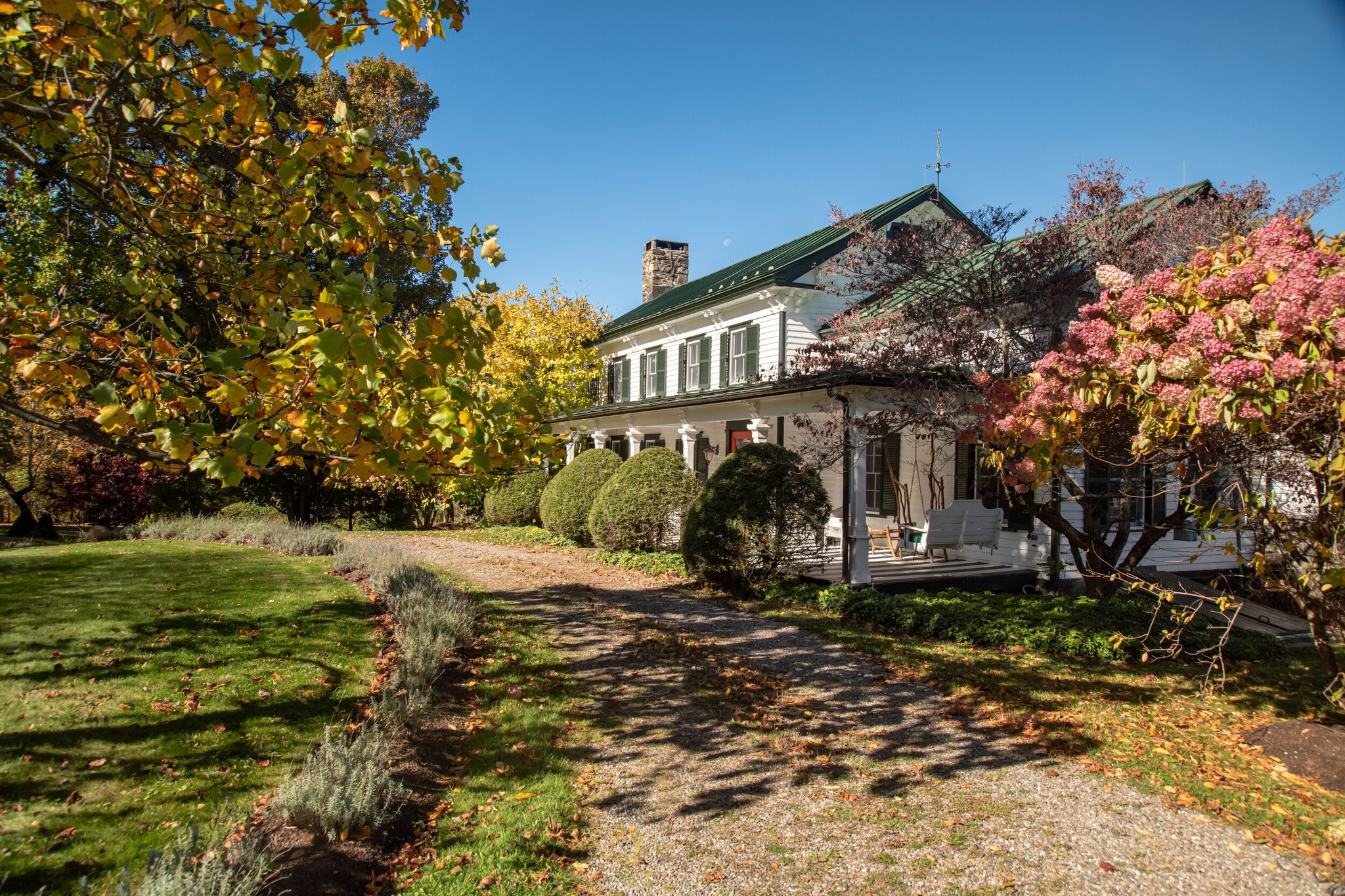 A gravel drive leads past lavender and pink hydrangea toward a porch-wrapped farmhouse in mid-October.