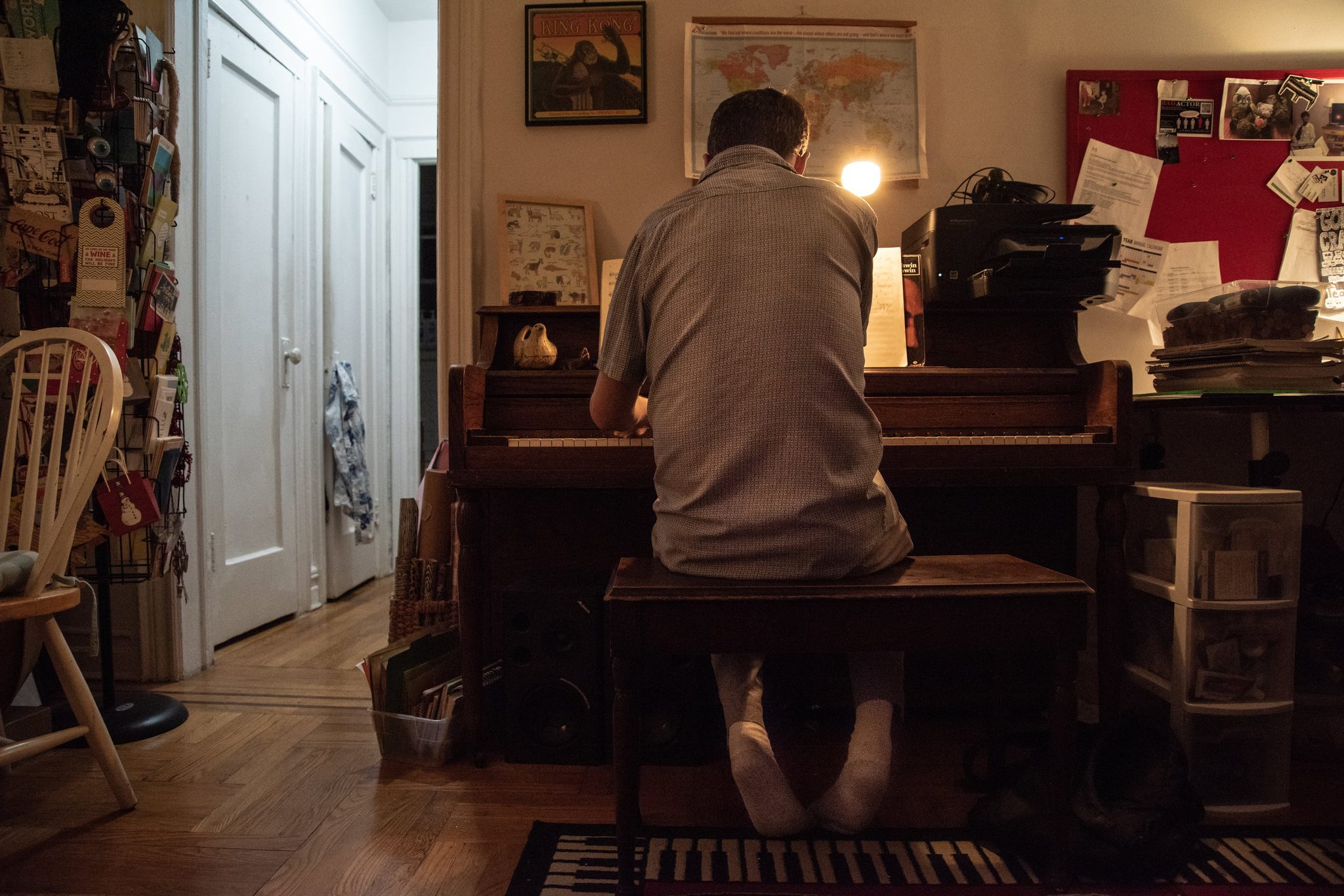 Tom works at the upright in the corner of the apartment, a map of the world pinned above.