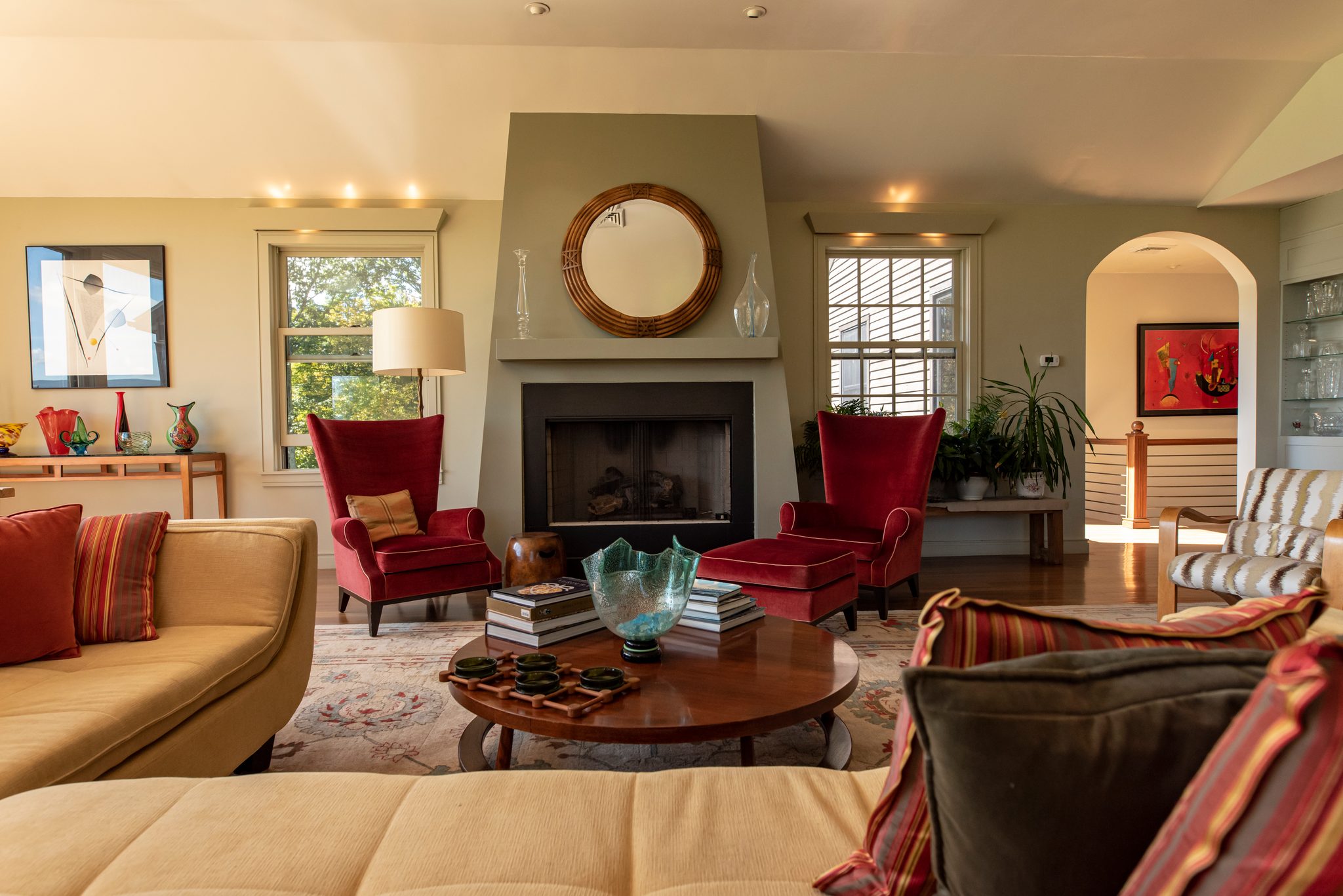 Red wing chairs and a round mirror anchor a bright living room, its fireplace flanked by tall late September windows.