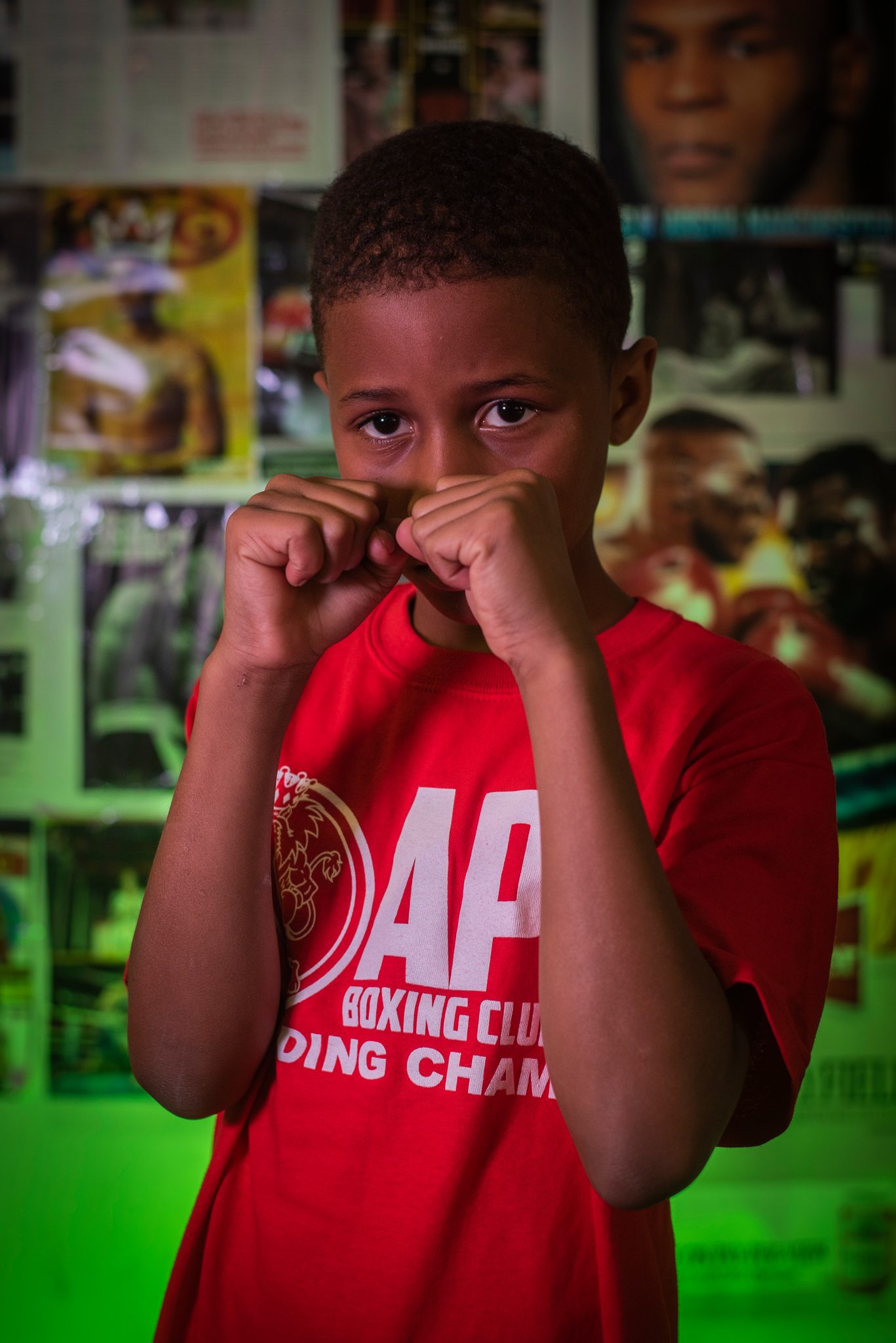 Peeking over his fists, a small boxer stands against the wall of fight posters and magazine covers.