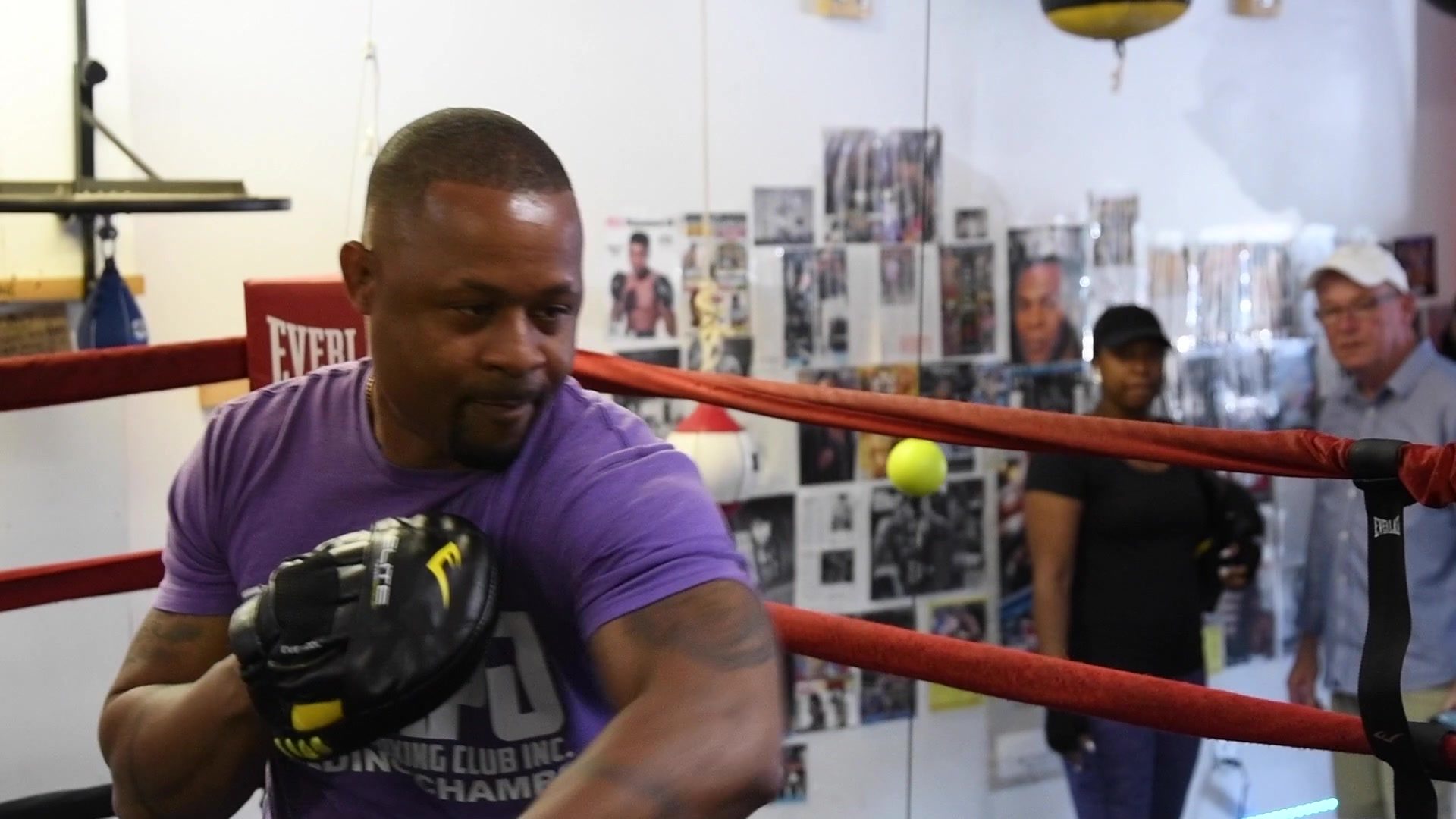 Coach Kariym Patterson, the club's founder, holds a mitt at the ropes in a purple club shirt, eyes on his boxer.