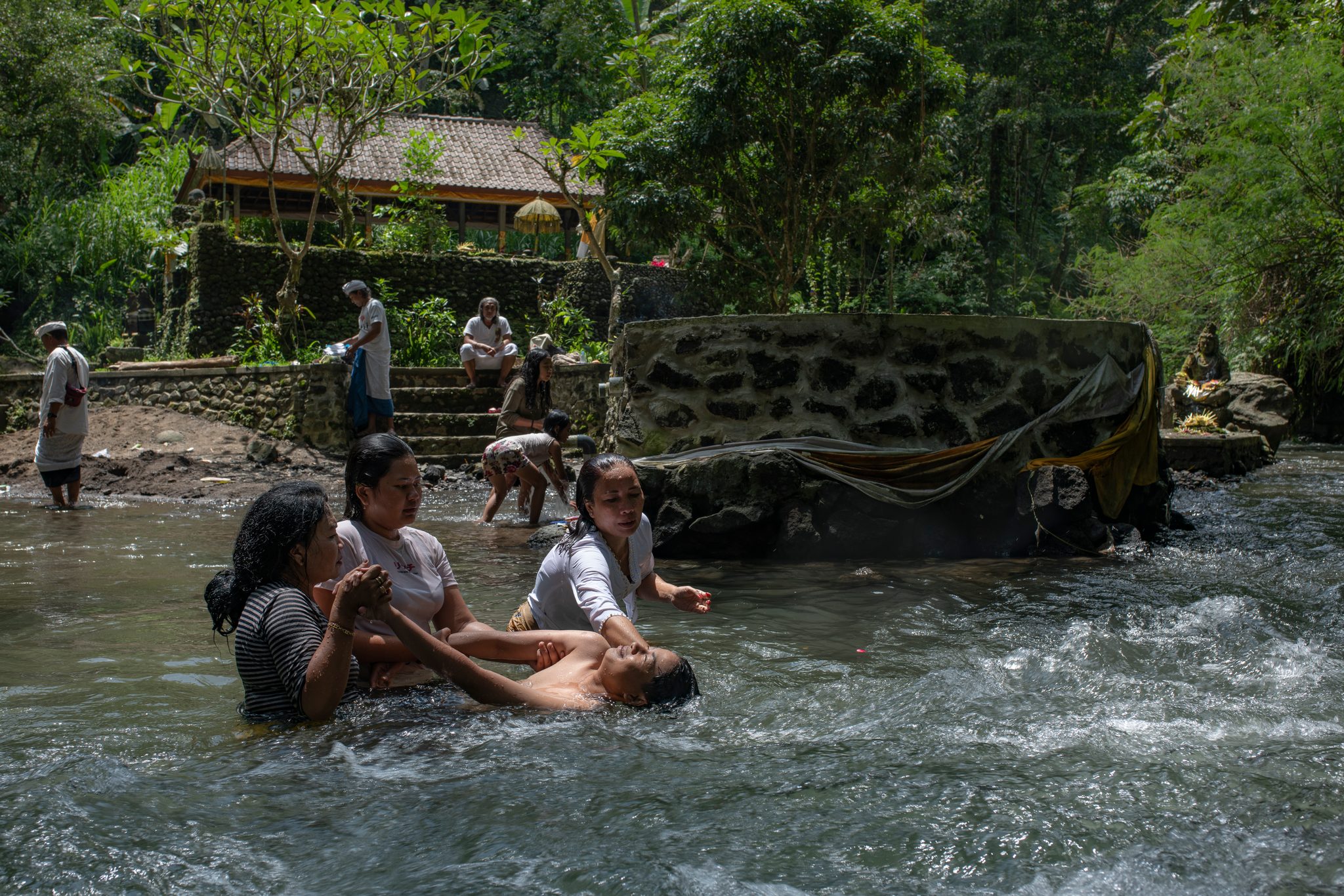 Families wade in the river below the temple, a man stretched out in the current.