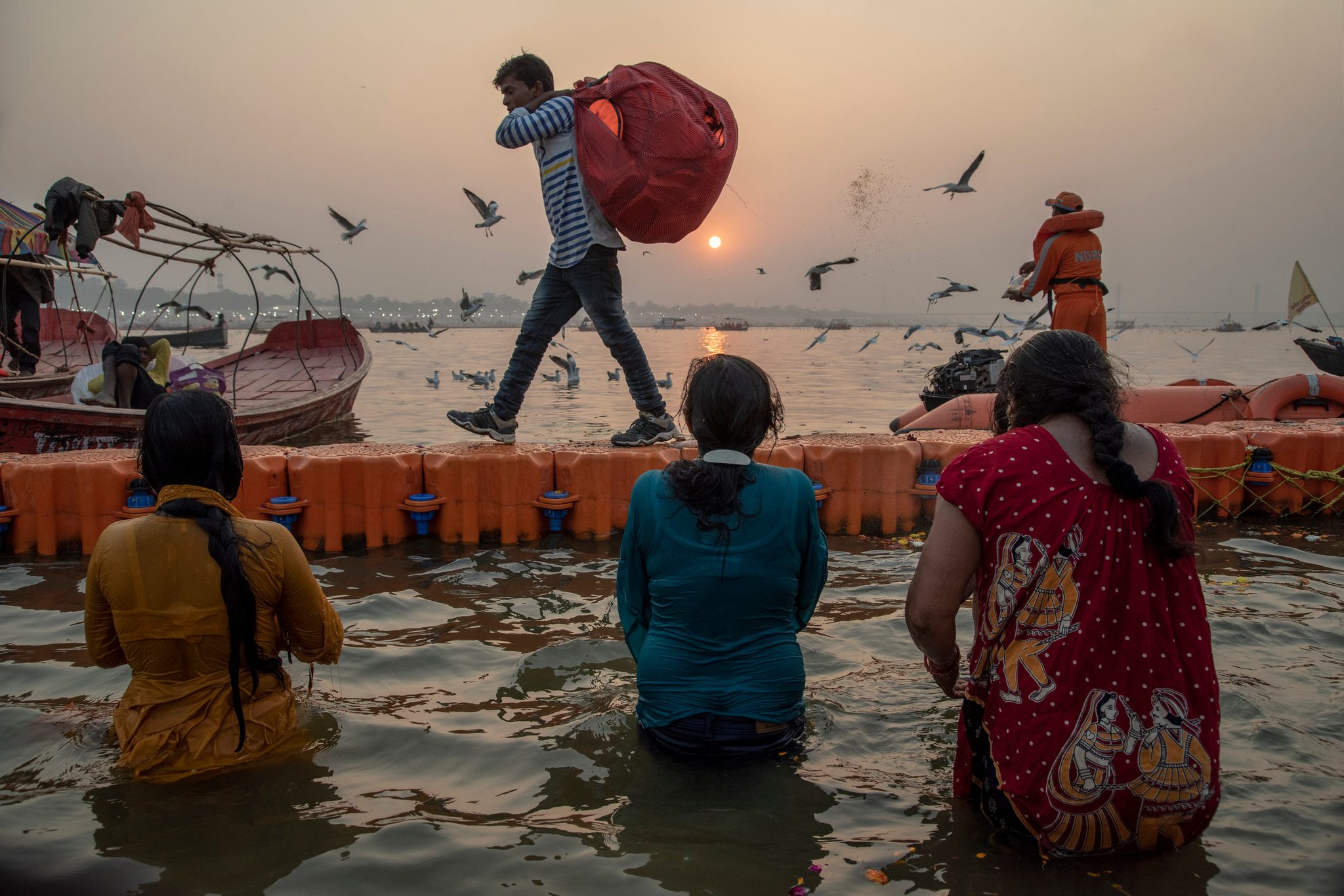 Three women bathe in the Ganges. A man carries life jackets along the barrier. Another tosses seed to the birds. Here at the rivers edge life gives and it receives. It preserves and purifies.