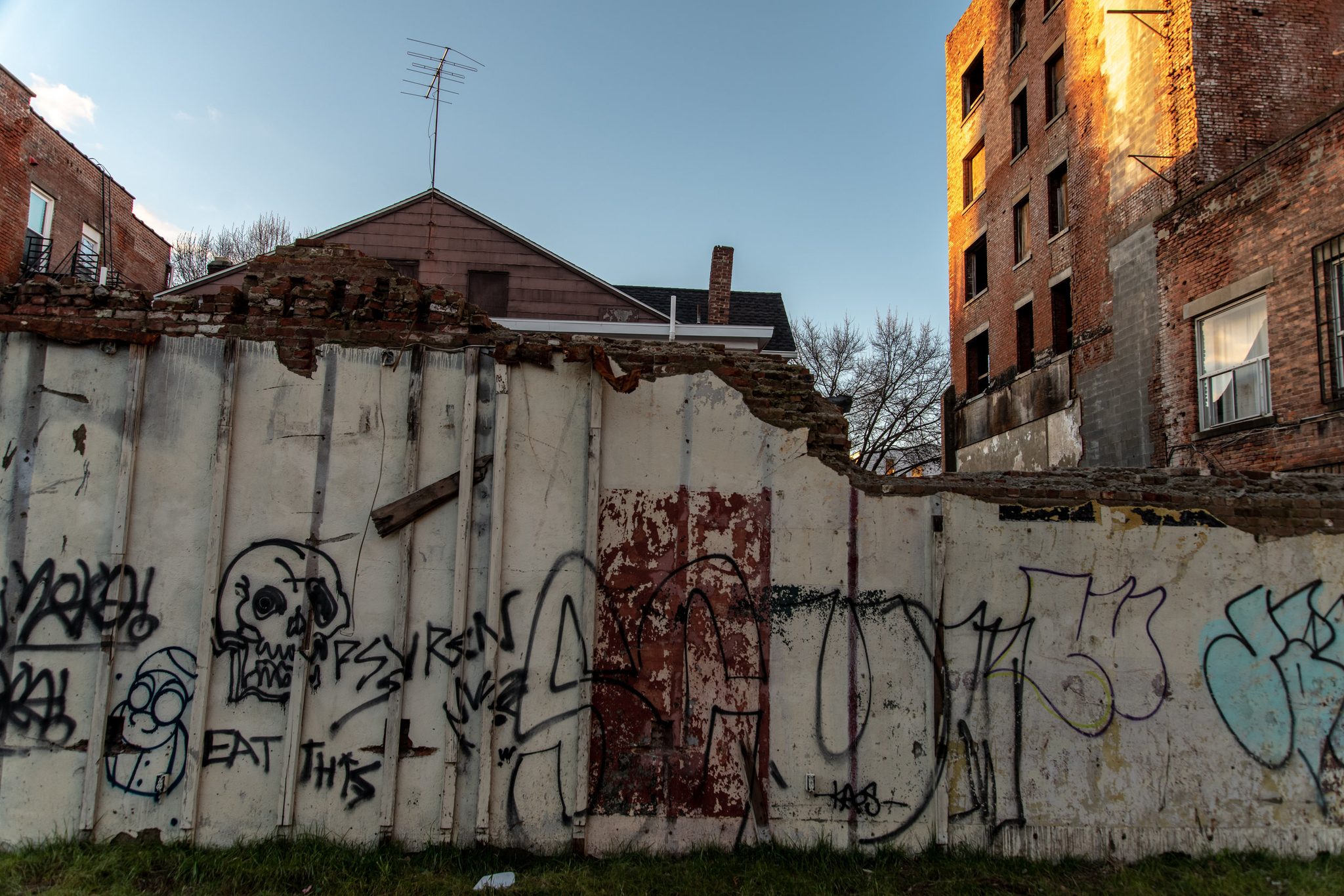 A graffiti-covered wall crumbles in front of an old brick building.