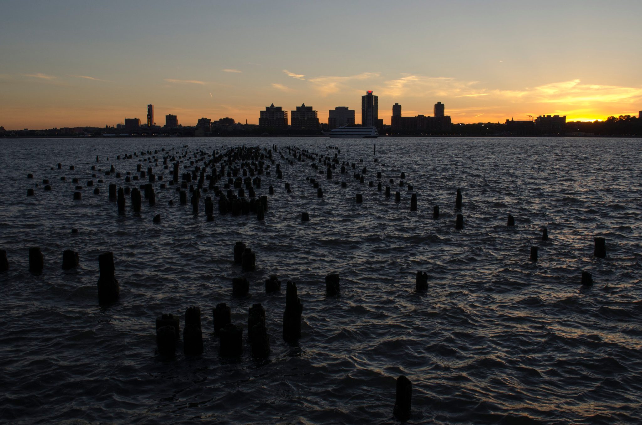 Old pier pilings rise from the Hudson like a forest of stumps as the sun goes down.