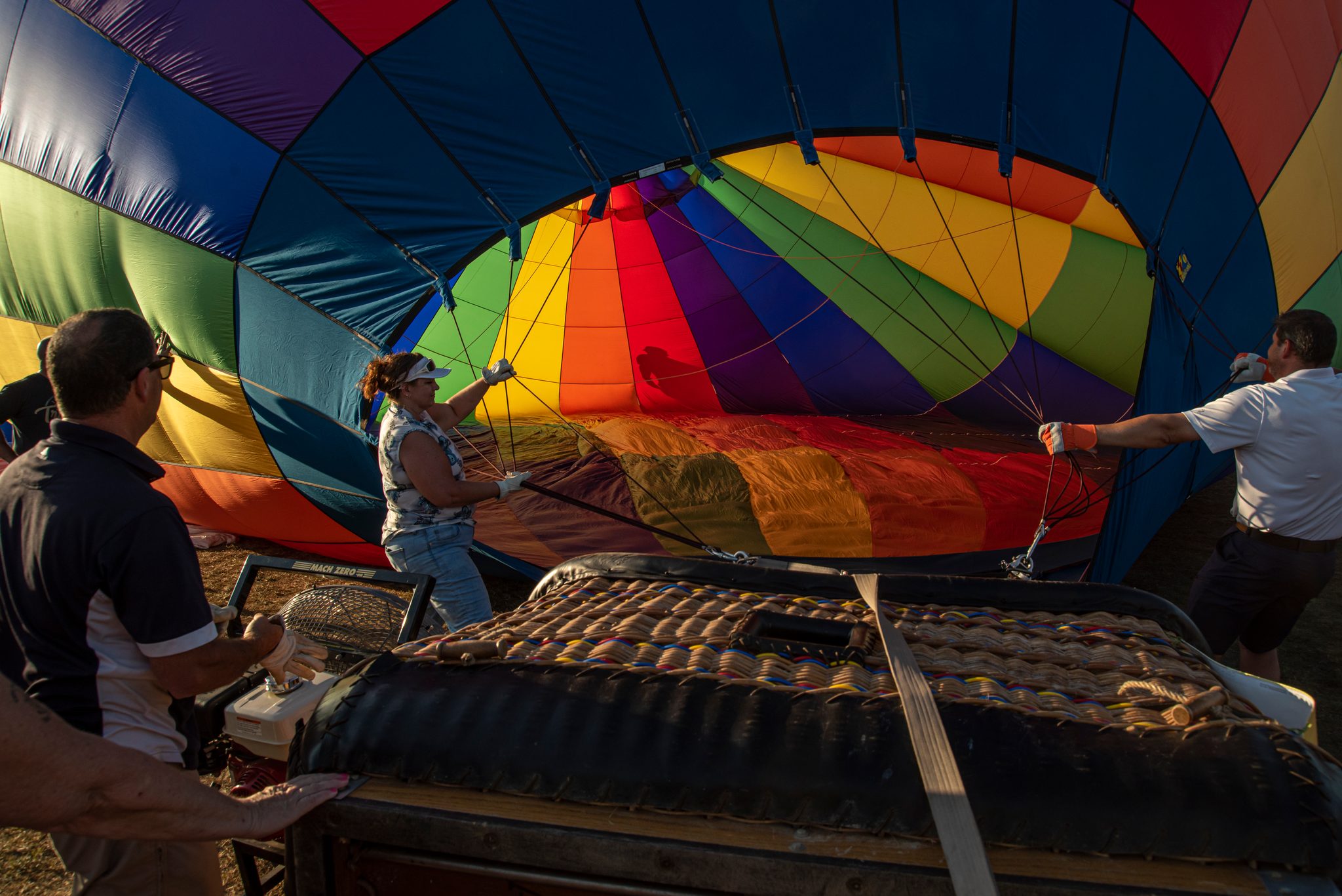 The crew holds open the mouth of a rainbow envelope while the basket waits on its side. Every flight begins like this, held up by many hands.