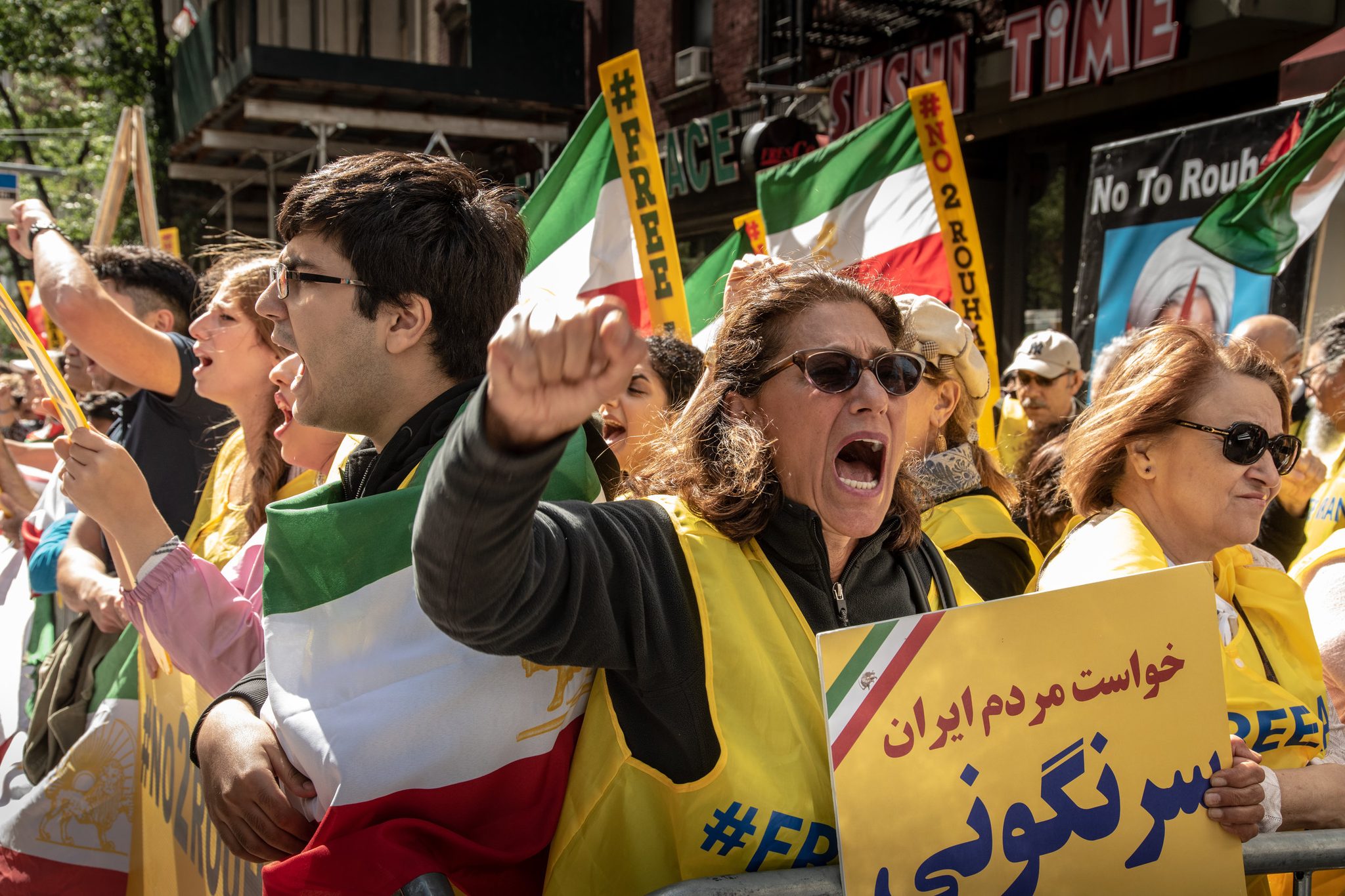 A woman shouts over a sign in Farsi as Iranian flags crowd the barricade. Beside her, a placard in English reads No to Rouhani.