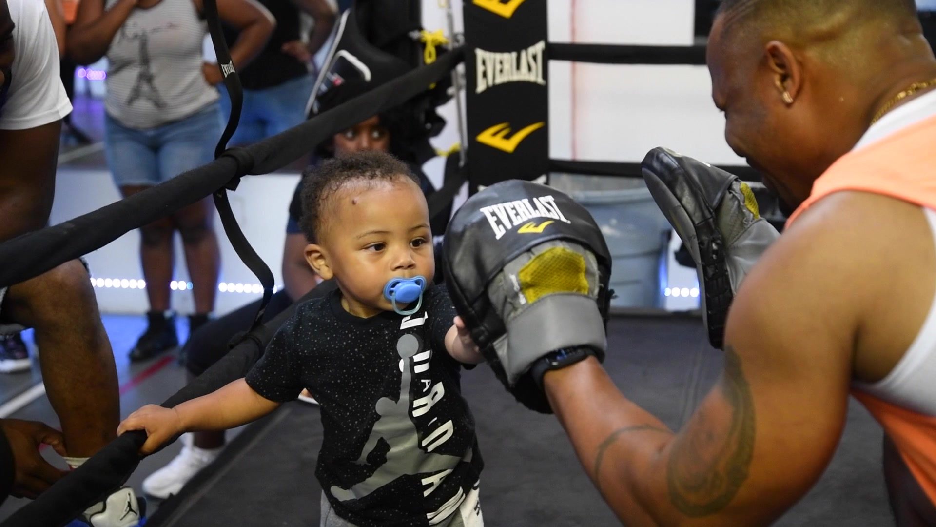 A toddler with a blue pacifier presses one hand into a coach's mitt, holding the bottom rope with the other.