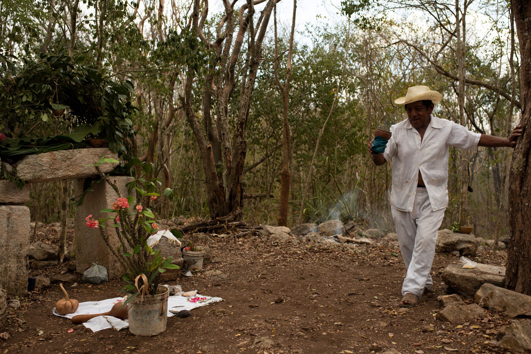 An elder in white and a straw hat walks up to the forest altar, a censer in hand.
