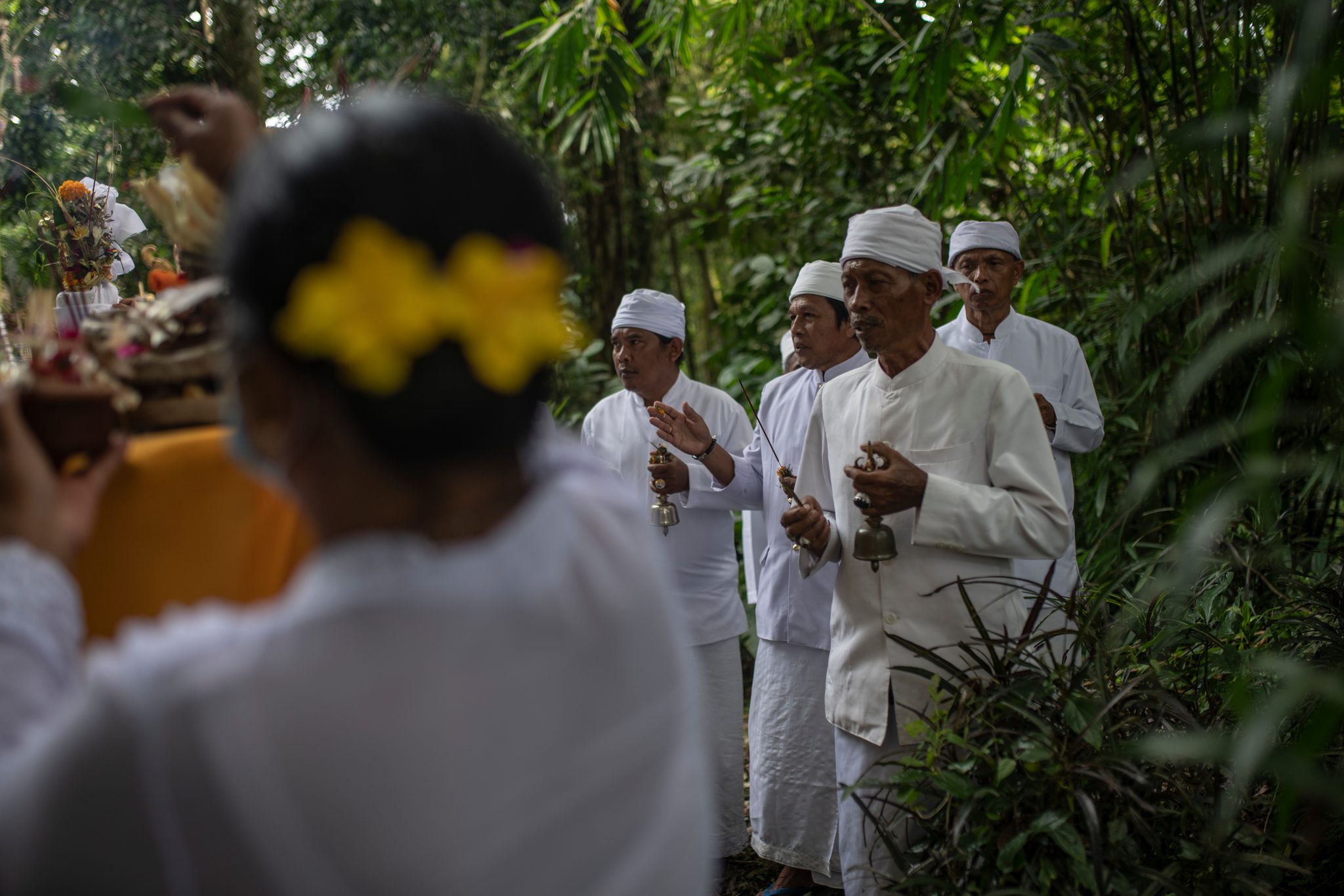 Priests ring their bells at the edge of the forest. A woman with yellow flowers in her hair carries an offering past.